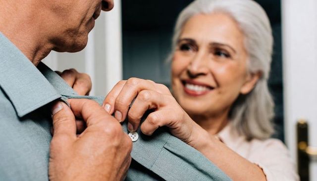 Senior couple sharing intimate caregiving moment fastening shirt collar at doorway