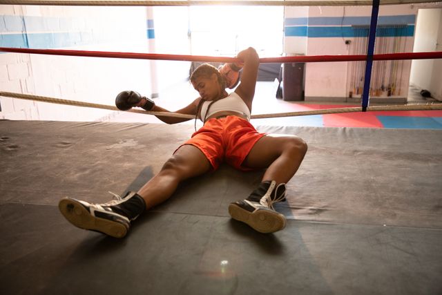 Depicts a biracial female boxer lying on the floor of a boxing ring, appearing knocked out. She is wearing sports clothes and boxing gloves. This image can be used to illustrate themes of strength, sports achievement, determination, and the physical demands of boxing. Suitable for articles, sports promotions, fitness blogs, and motivational content.