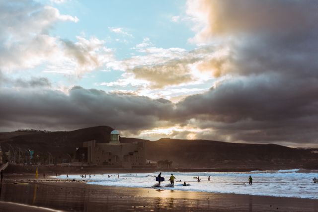 Surfers ride waves against a dramatic cloudy sunset sky on a scenic beach. Mountains surround the coastline adding to the natural beauty. Ideal for promoting beach activities, water sports, travel destinations, and adventure vacations.