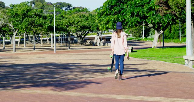 A teenage girl in casual attire with a blue baseball cap, walking on a park path while carrying a skateboard. Trees and greenery can be seen around, creating a peaceful urban environment. Great for themes related to youth lifestyle, outdoor recreation, and casual day activities.