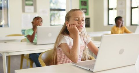 Bored blonde girl using laptop while African classmates studying in bright classroom, pensive studen