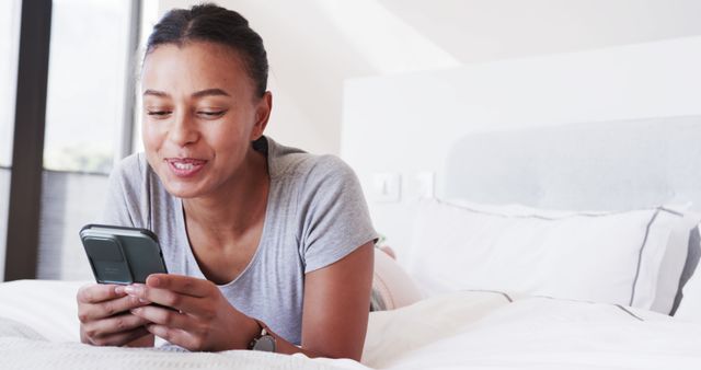 Woman lying on comfortable bed using her smartphone with a smile. Great for promoting mobile apps, lifestyle blogs, social media content, technology advertisements, or health and wellness articles.