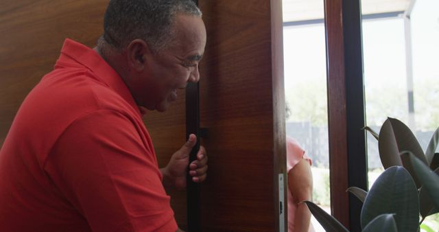 Friendly middle-aged man in red shirt happily opening front door to greet visitor. Green indoor plant accents visible, adding warmth to home entrance. Perfect for concepts like greeting, hospitality, home visits, and friendly interactions.