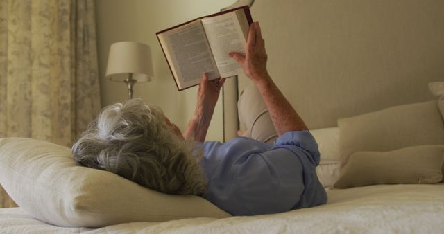 An elderly woman is seen lying on a comfortable bed, reading a book. This image can be used to highlight themes of relaxation, leisure, and senior lifestyle. Suitable for articles or advertisements related to aging, home comfort products, and mental well-being among the elderly.