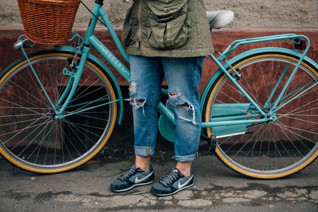 Person standing next to vintage teal bicycle with basket on an urban street. Wearing ripped jeans and casual shoes contributes to relaxed, trendy city vibe. Perfect for use in urban lifestyle, fashion, apparel, travel, and leisure themes.