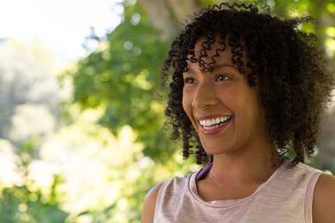 Joyful woman smiling in sunlit park with curly hair and casual sleeveless top portrait