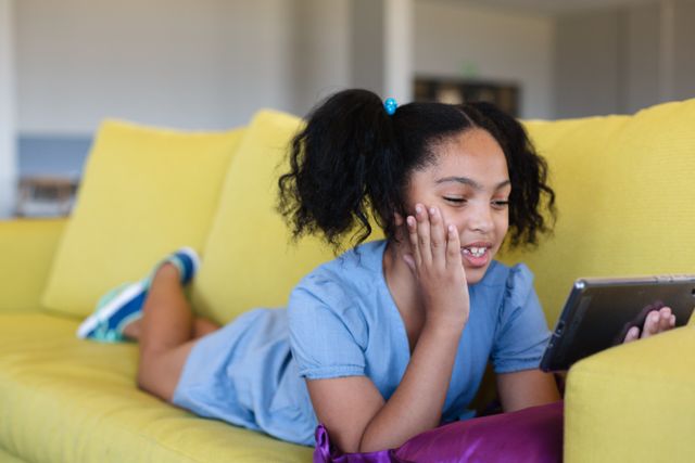 Smiling biracial elementary schoolgirl touching cheeks while using digital tablet on couch in school. unaltered, childhood, education, learning, wireless technology, relaxation and school concept.