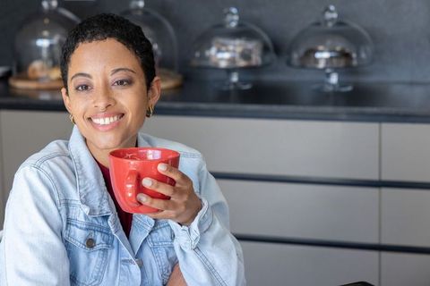Smiling african american woman holding red mug at modern kitchen counter in denim jacket
