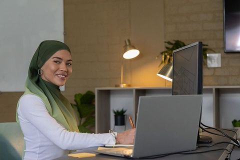 Woman wearing green hijab working at office desk with laptop and external monitor