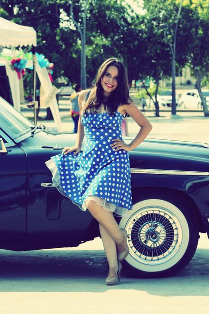 Woman wearing a blue polka dot dress posing next to a vintage car outdoors. Trees and park setting in the background suggest a warm summer day. Perfect for use in fashion editorials, retro style promotions, summer event marketing materials, and automotive advertisements.