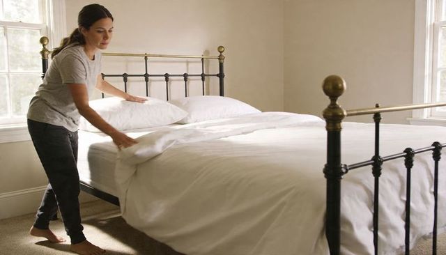 Woman making bed in sunlit minimal bedroom with white duvet and brass metal bedframe
