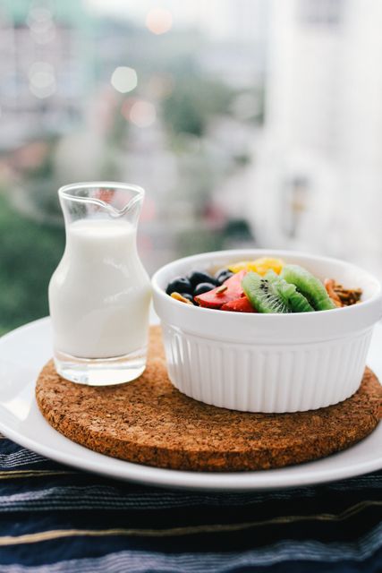 Delicious and colorful breakfast bowl placed on a white plate includes fresh fruits like kiwi, strawberry, and blueberry. Beside the bowl is a small glass bottle of milk. Ideal for promoting healthy eating, nutrition blogs, breakfast recipes, and dairy products.