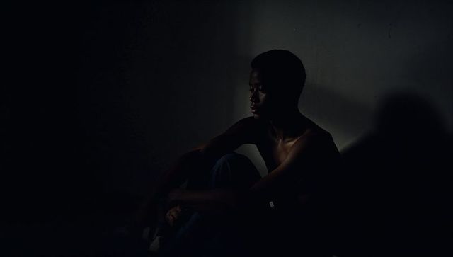 Moody low-key portrait of shirtless teen hugging knees in dim room with spotlight