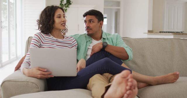 Couple in casual clothes enjoying time together on a comfortable couch in their living room. They are using a laptop and sharing a joyful conversation. Ideal for ads related to home lifestyle, relationship tips, technology products, or family bonding themes.