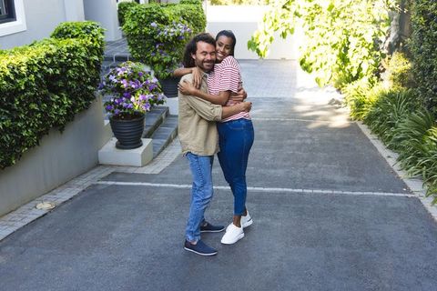 Smiling interracial couple hugging on sunny driveway with lush hedges and planters