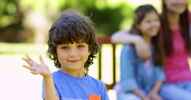 Curly-haired boy smiling and waving with family in background. Capturing moments of joy and togetherness in an outdoor setting. Ideal for promoting family activities, outdoor events, and child-friendly products.