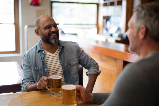 Two men are sitting at a restaurant, enjoying glasses of beer and engaging in conversation. They are smiling and appear to be having a good time. This image can be used for themes related to friendship, socializing, leisure activities, and casual dining experiences.