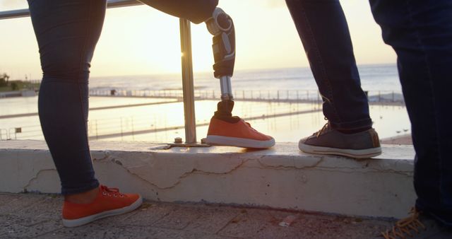 Two people standing near ocean at sunset, one with a prosthetic leg, capturing a moment of support and determination. Great for use in topics related to inclusivity, resilience, and adaptive sports.