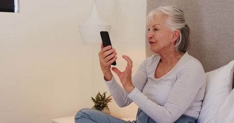 Senior woman video calling on smartphone while relaxing in bedroom