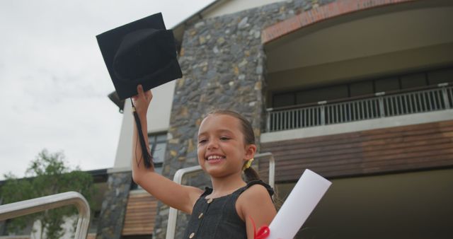 Young girl celebrating graduation by holding up cap and diploma in front of modern school building. Bright, happy expression conveys pride and achievement. Ideal for educational themes, milestone celebrations, graduation announcements, and school-related promotions.