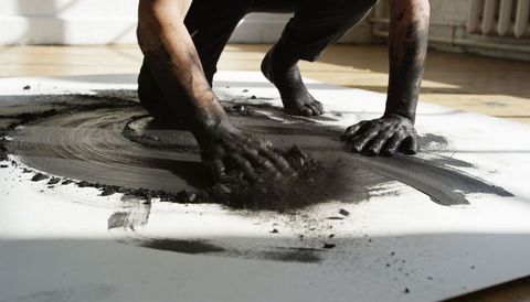 Artist kneeling and spreading black pigment with hands on white canvas, charcoal motion