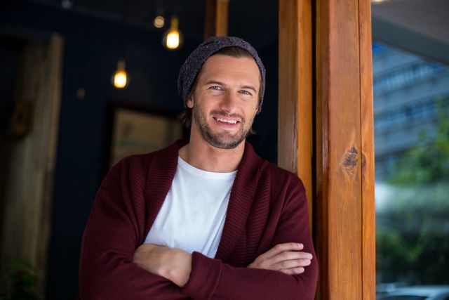 Man leaning against window in a café, wearing a beanie and casual clothing. Ideal for use in lifestyle blogs, urban living articles, or advertisements promoting casual fashion and relaxed environments.