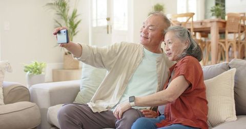 Senior asian couple taking selfie on sofa in sunlit living room with smartwatch