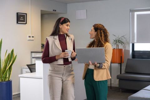 Diverse female coworkers collaborating in modern office lounge holding laptop and coffee
