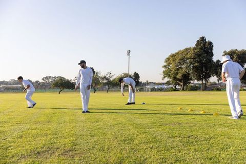 Diverse male cricket team practicing fielding drills on sunlit green park pitch