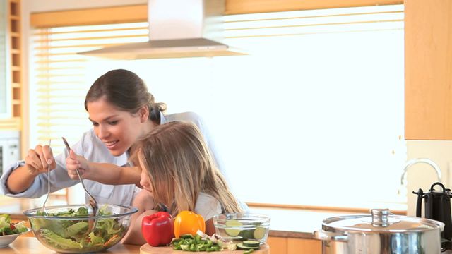 A mother and daughter enjoy quality time preparing a fresh salad together in a modern, bright kitchen. The setting promotes wholesome eating and family bonding. Perfect for use in marketing materials for culinary courses, healthy lifestyle blogs, parenting tips, kitchen appliance advertisements, and family-oriented publications.