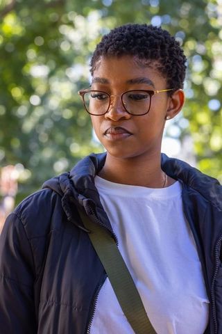 Thoughtful young woman wearing round eyeglasses and puffer jacket in sunlit city park