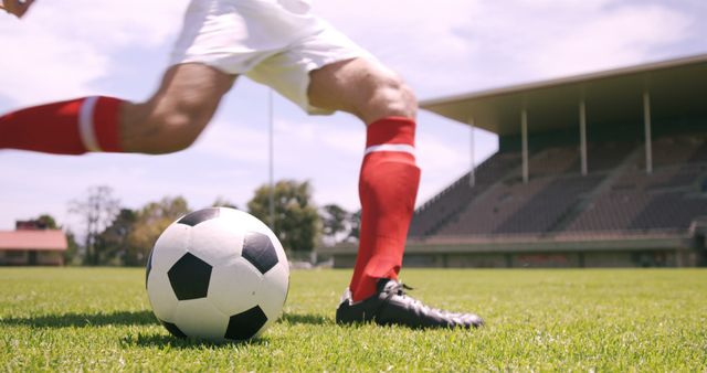 Close-up of a soccer player mid-action as they kick the ball on a grassy field. The player wears red socks and black shoes, showcasing dynamic movement and intensity. Ideal for illustrating sports articles, athletic gear advertisements, and team practice flyers.