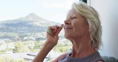 Senior woman using jade roller while relaxing by window with mountain view