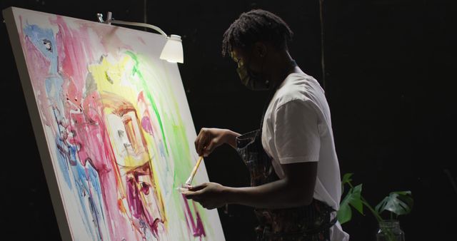 An artist is painting on a large canvas, illuminated by a lamp, in a dark studio. This image can be used to depict creativity, artistic processes, or safety measures during the pandemic. Ideal for articles or blogs about art, creativity workshops, or the impact of COVID-19 on artists.