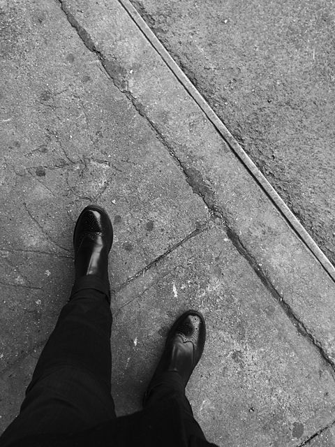 Photo taken from a first-person perspective showing a person walking on a city sidewalk. The shoes are polished and stylish, adding a touch of fashion to an everyday urban scene. Perfect for use in articles about city life, urban fashion, street photography, and personal blogs discussing daily activities or fashion choices.
