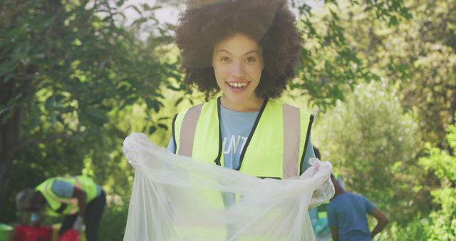 Young woman wearing yellow safety vest smiling while holding empty trash bag during park clean-up. Other volunteers picking up litter in background. Use for promoting community service, environmental awareness, outdoor activities, teamwork, and volunteer programs.