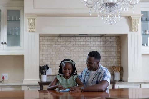 African american father teaching daughter on tablet at kitchen island under chandelier
