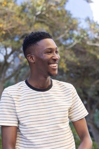 Smiling young man enjoying sunny park moment while wearing striped knit t-shirt in daylight