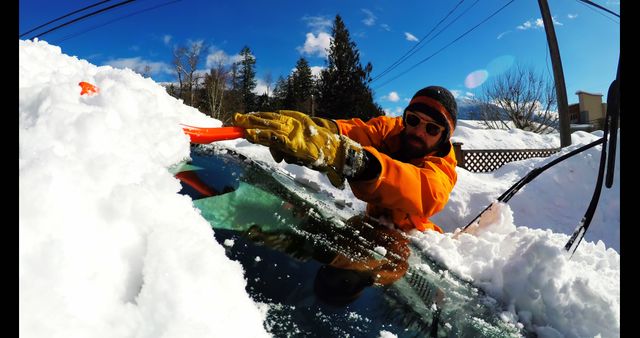 Man wearing orange jacket and gloves cleaning snow off car windshield with a snow brush on a bright sunny winter day. Useful for themes related to winter weather conditions, car maintenance, snow removal, winter safety, road trip preparations, and outdoor winter activities.