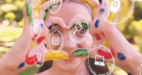 Holding painted hands girl framing eyes with heart gesture, floating digital learning icons