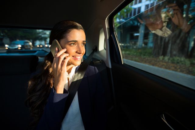 Businesswoman talking on mobile phone while traveling in car, smiling and looking out window. Ideal for illustrating business travel, professional communication, commuting, and modern work lifestyle.
