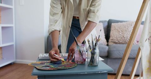 Artist mixing paint on well-worn palette with brushes in jars on home studio table