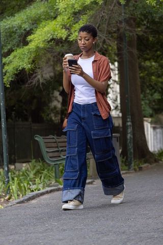 African woman walking in park checking smartphone and holding takeaway coffee