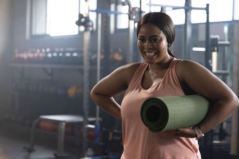 Smiling african american woman holding green yoga mat in gym sunlit fitness moment