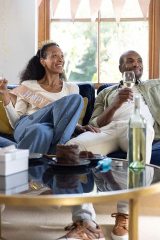 Couple celebrating at home wearing happy sash and tiara while holding wine and cake