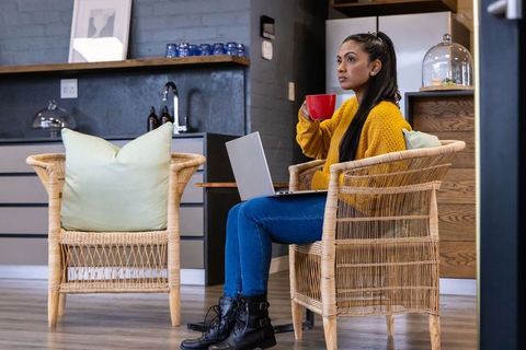 Asian woman working from home in cozy kitchen cafe holding laptop and red mug