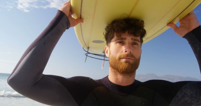 A young Caucasian man in a wetsuit holds a surfboard on a sunny beach, with copy space. His confident expression and the clear sky suggest an enjoyable day of surfing ahead.