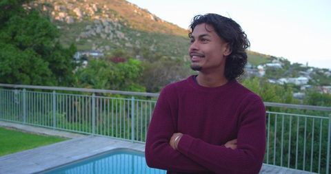 Young man standing on pool deck beside pool overlooking sunlit hillside in maroon sweater