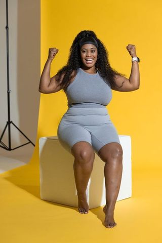 African american woman flexing and smiling in gray activewear with smartwatch on yellow backdrop