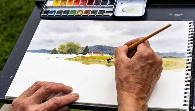 Senior artist painting plein air watercolor landscape in spiral sketchbook with palette